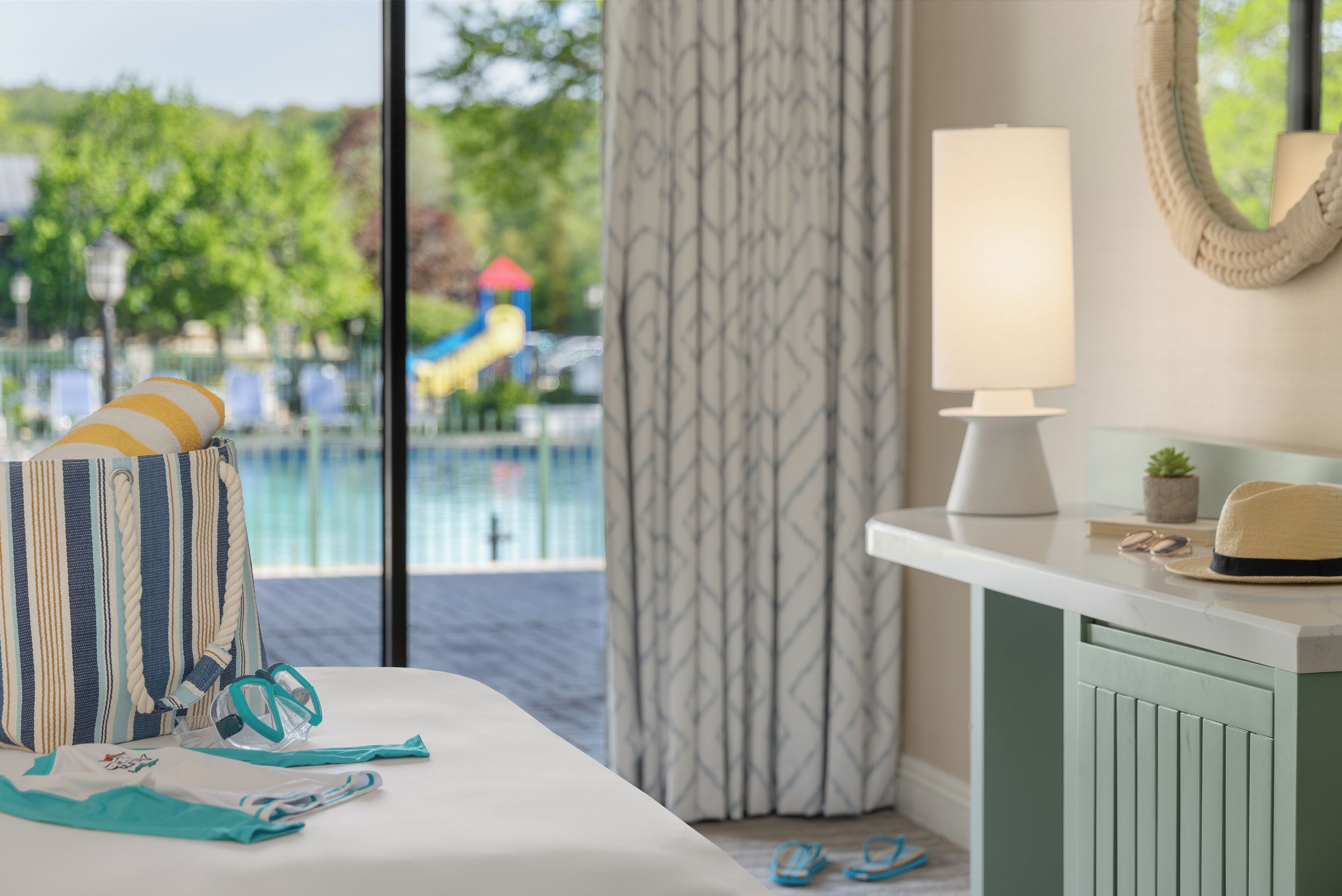 A hotel room with a bed, a bedside table with a lamp, a suitcase, and a view of a pool and playground.