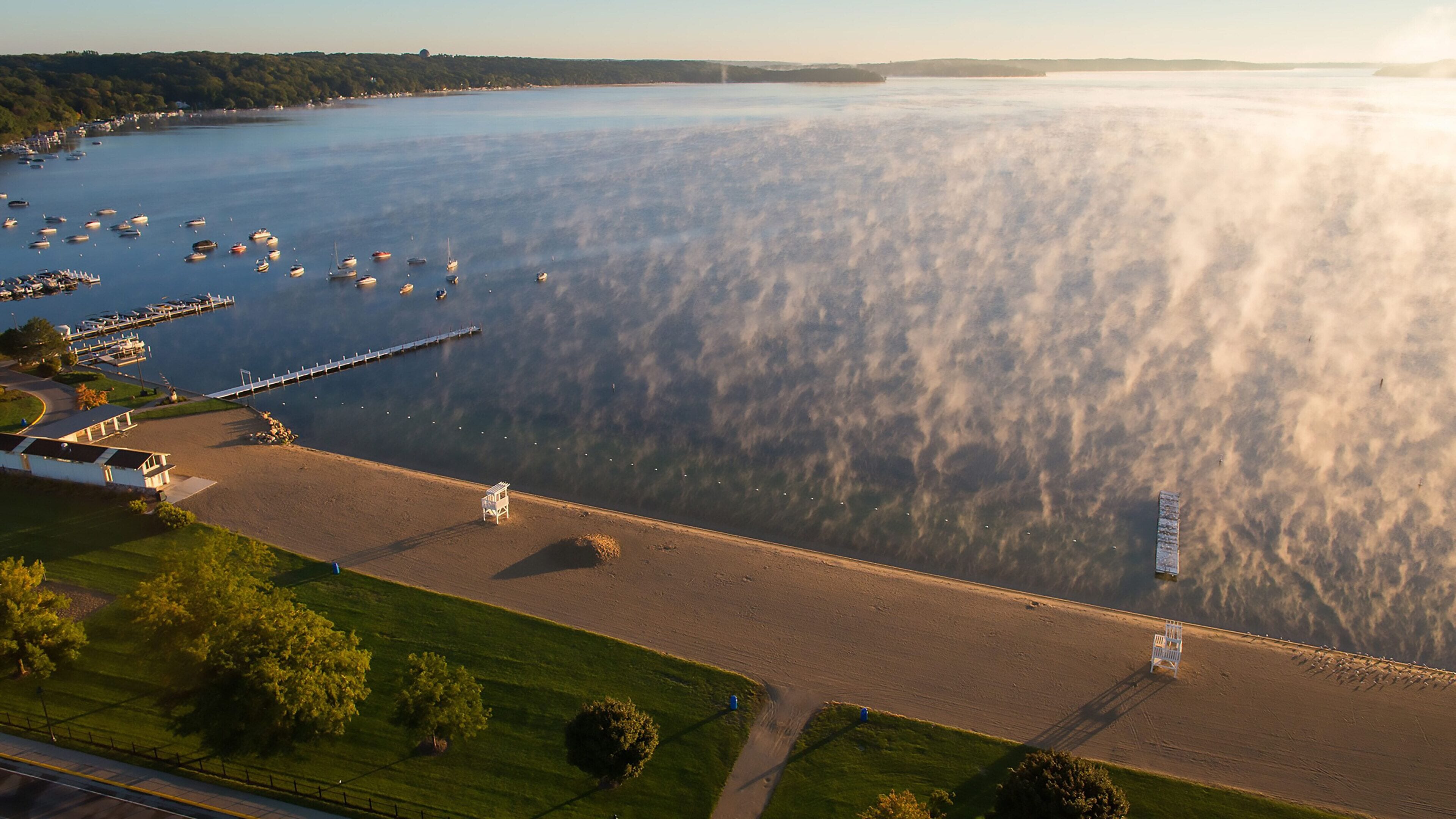 Vue aérienne d’une zone côtière comprenant un port de plaisance, une plage de sable et une zone herbeuse avec des arbres.