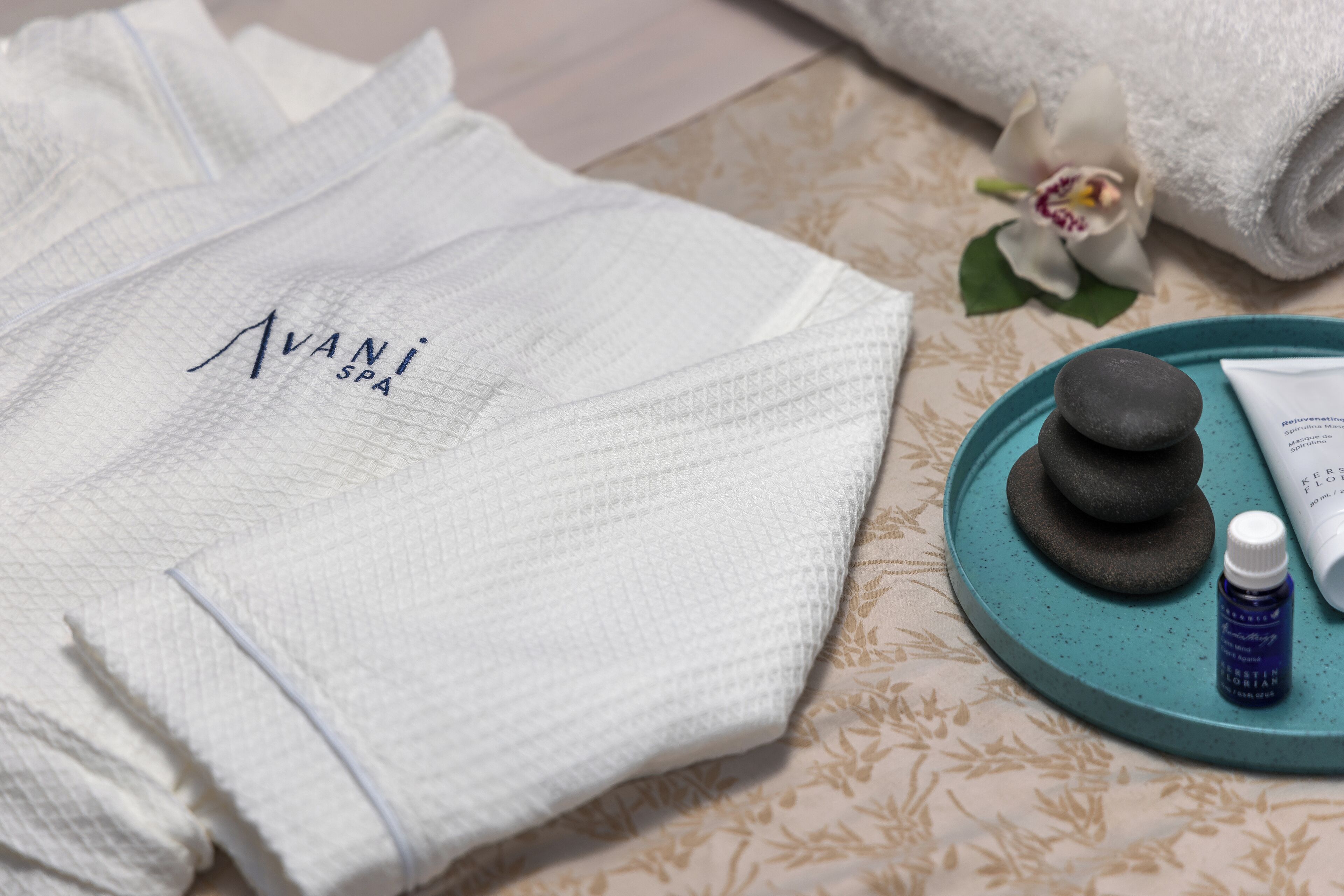 Una toalla blanca con el texto "AVANI SPA", una herramienta de masaje de piedra y una botella sobre una bandeja color turquesa.