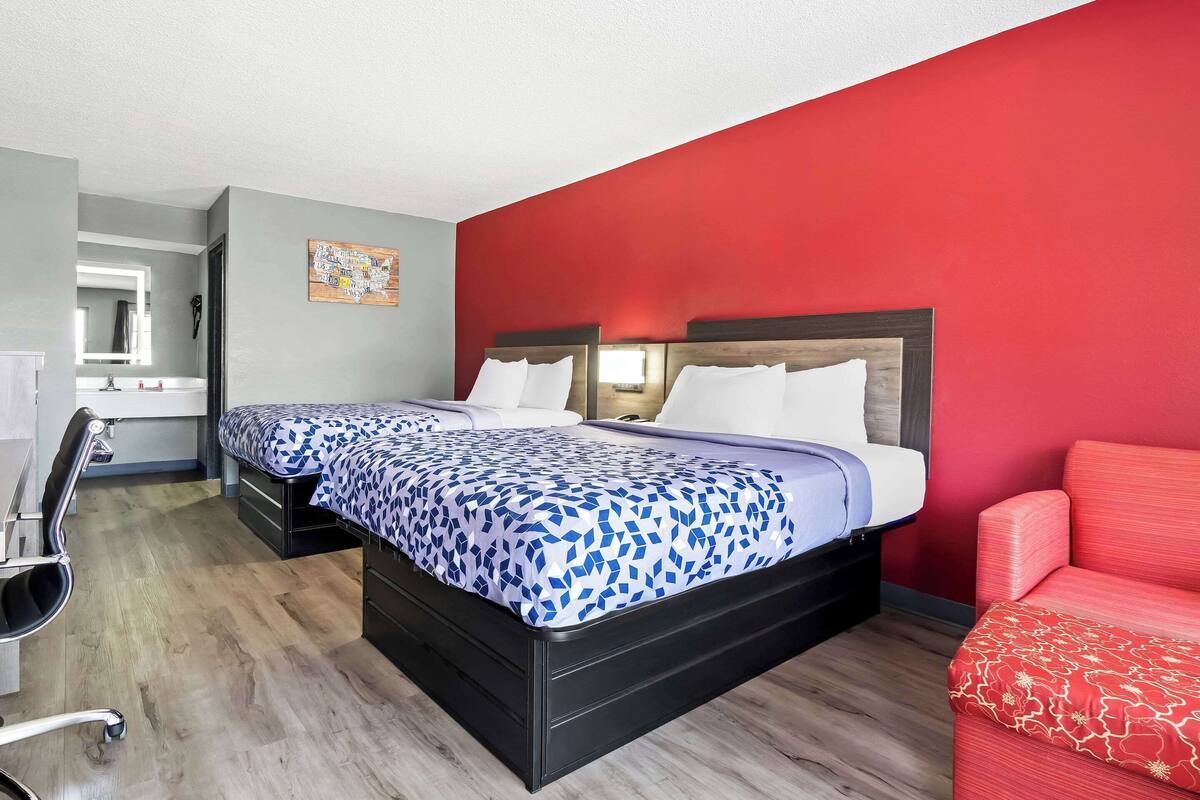 A hotel room with a double bed, a red accent wall, a desk with a chair, and a red sofa.