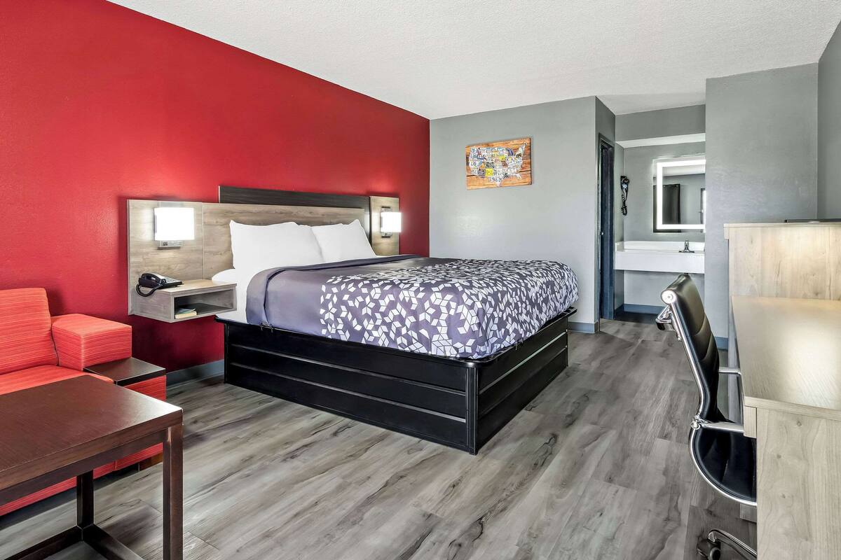 A modern hotel room with a bed, a red sofa, a wooden table, and a bathroom visible through an open door.