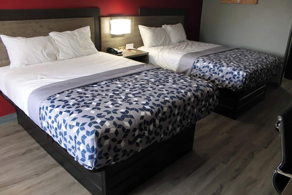 Two beds with white linens and patterned bedspreads, a bedside table with a lamp, and a wooden floor.