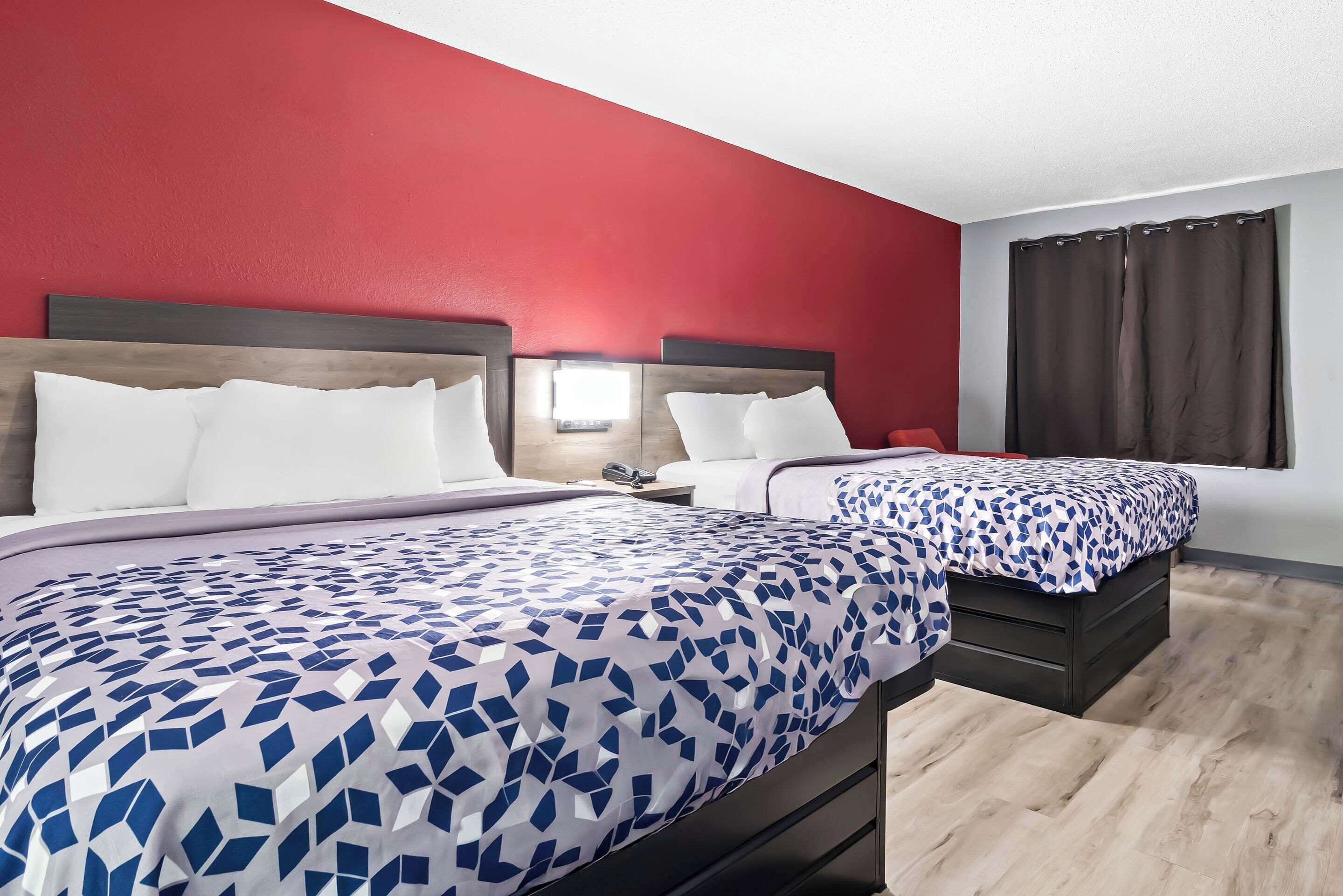 Deux lits avec des têtes de lit, un mur rouge, un plancher en bois et une fenêtre avec des rideaux.