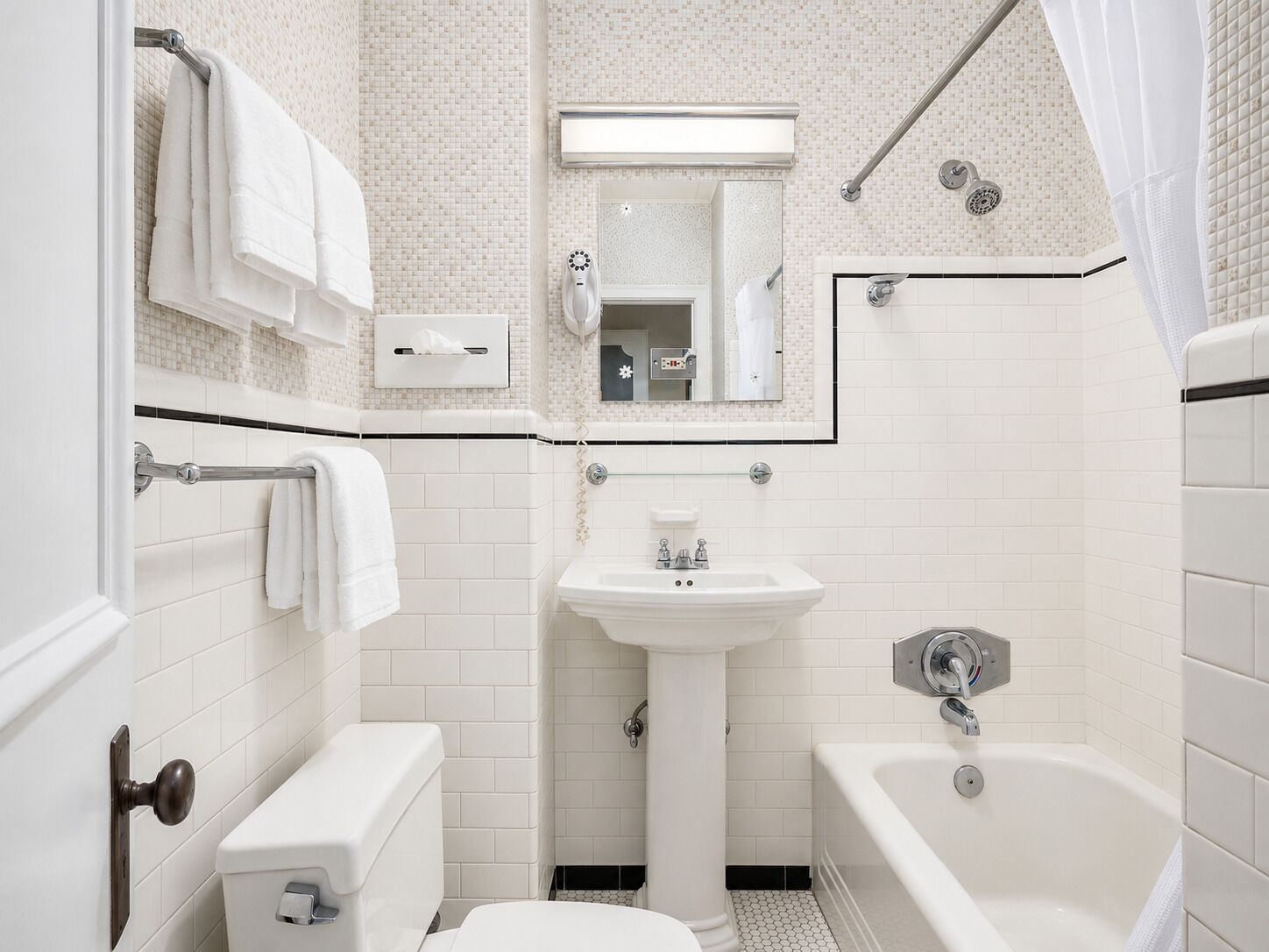 Et badeværelse med toilet, vask og badekar, alt i hvidt, med flisebeklædt bagvæg og et spejl over vasken.