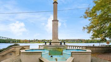 A monument with a fountain in the center, surrounded by a paved area with a stone wall and a bridge in the background.