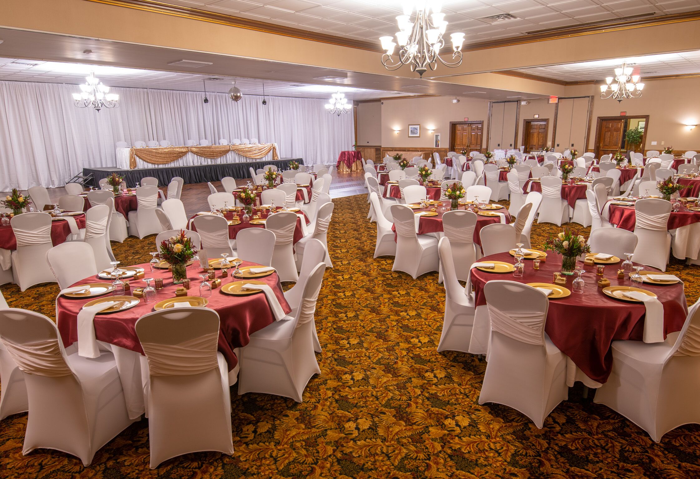 Un salón de banquetes con mesas redondas, sillas blancas y manteles rojos.