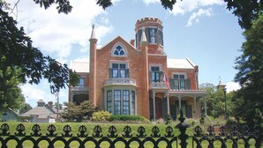 Un edificio grande y histórico de ladrillo con una torre y ventanas salientes, rodeado de un césped cuidado y una valla de hierro decorativa.
