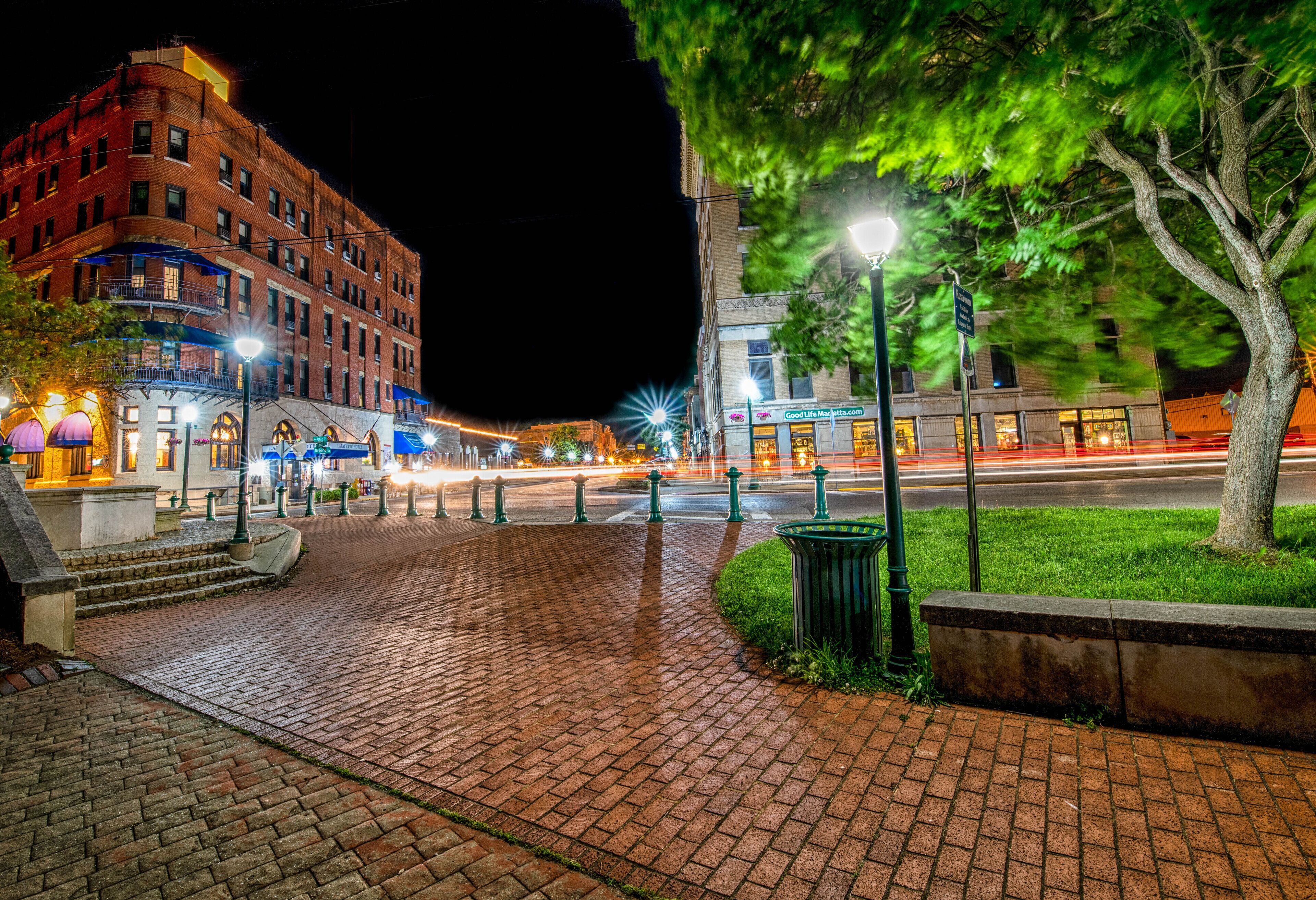 A nighttime city square with a brick pathway, street lamps, and buildings with illuminated signs.