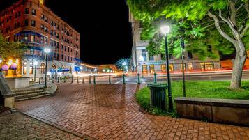 A nighttime city square with a brick pathway, street lamps, and buildings with illuminated signs.