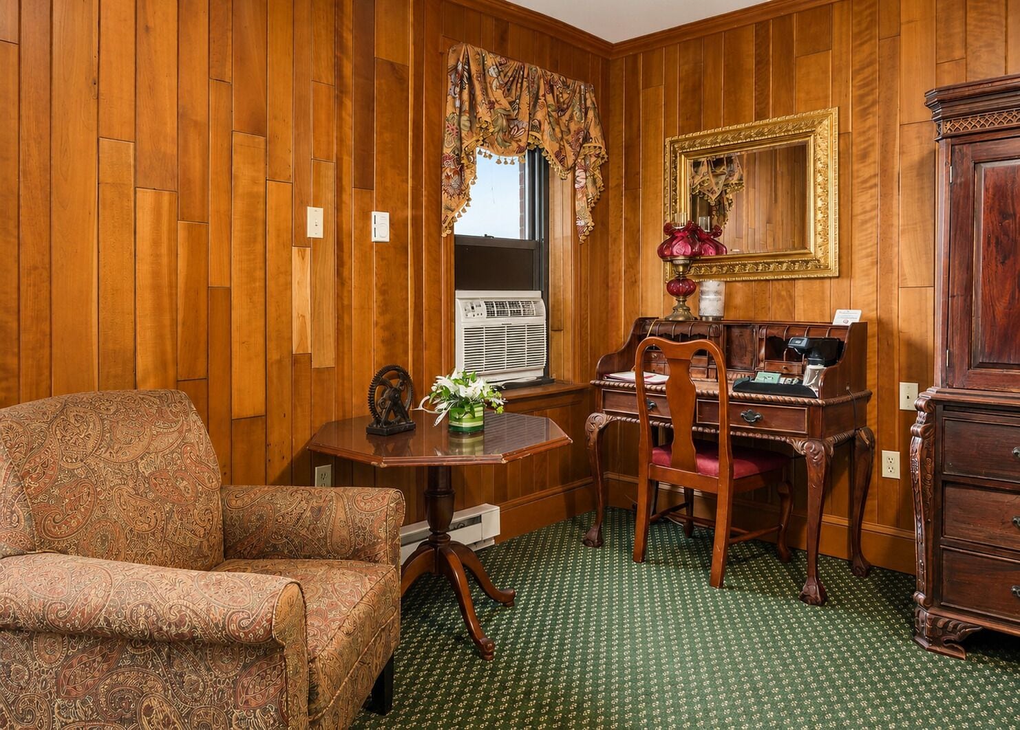 Una habitación con paneles de madera, un sillón estampado, un escritorio con lámpara, un espejo y un aparador.
