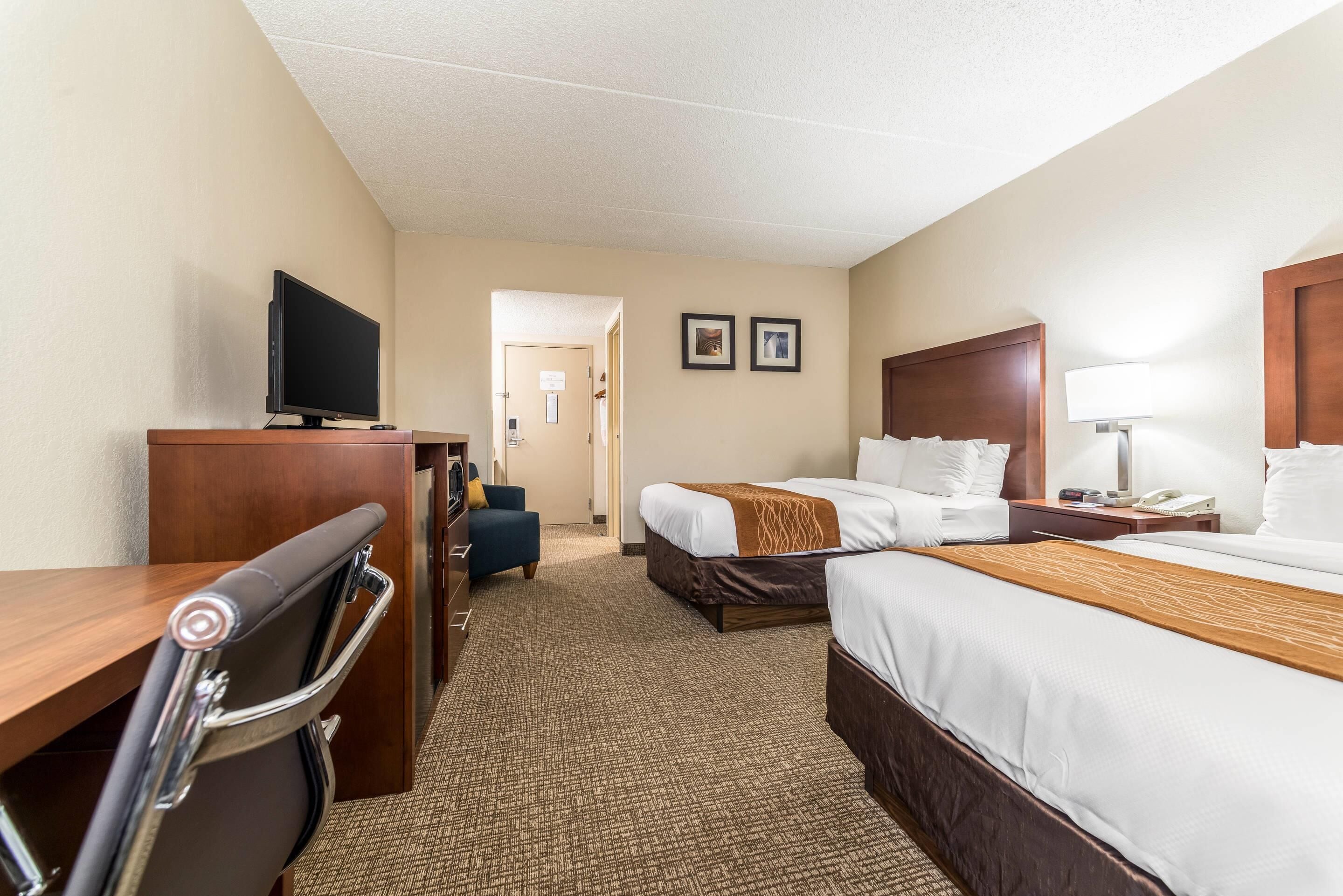 A hotel room with a desk, two beds, a TV, a chair, and a lamp.