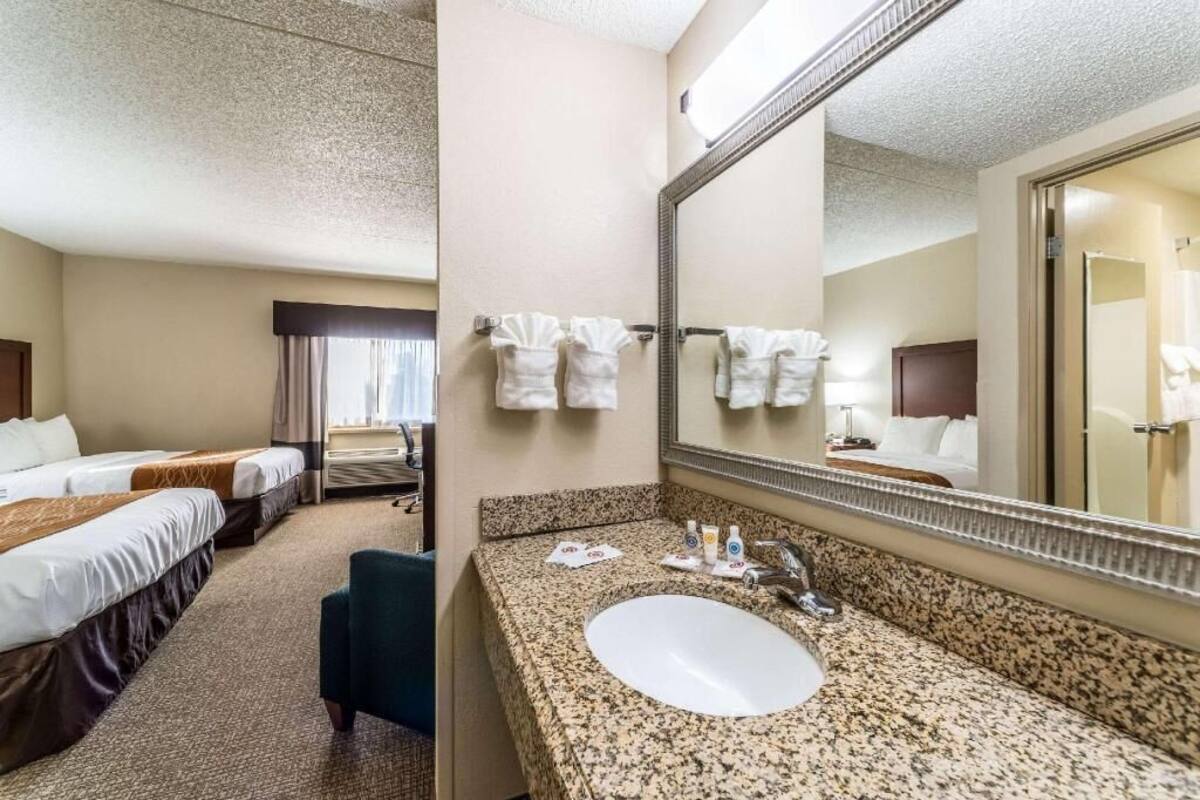 A hotel room with a bed, a sitting area, a bathroom with a sink and mirror, and a large window.