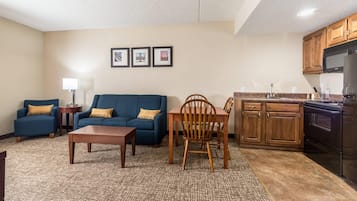 Una sala de estar con un sofá azul, una silla de madera, una mesa de centro y una cocina con armarios de madera y un refrigerador negro.