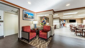 A hotel lobby with a reception desk, two red armchairs, a coffee table, a floral centerpiece, and a dining area with wooden tables and chairs. - Comfort Inn Festus - St Louis South (Festus)