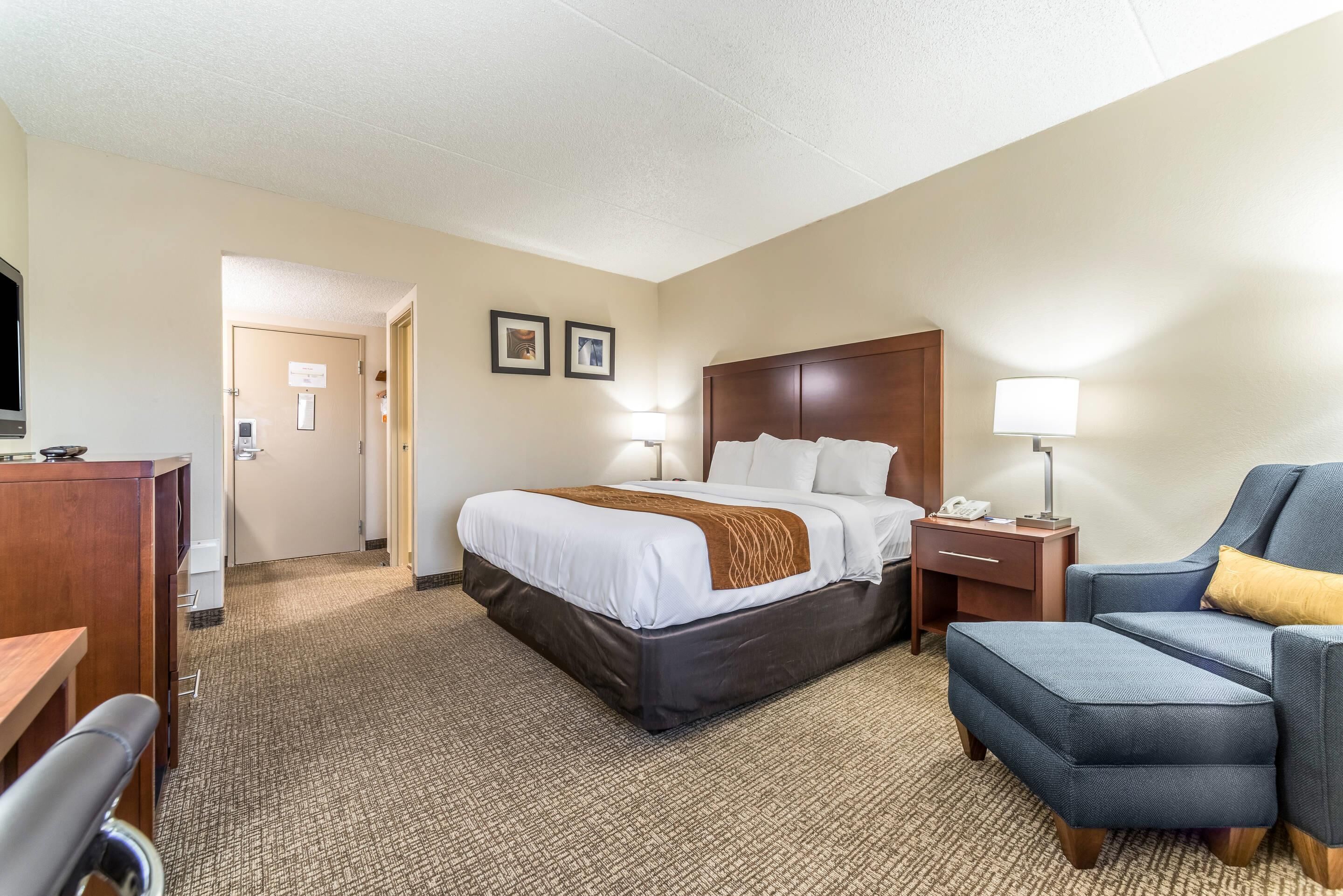 A hotel room with a bed, a desk, a chair, a nightstand, and a lamp.
