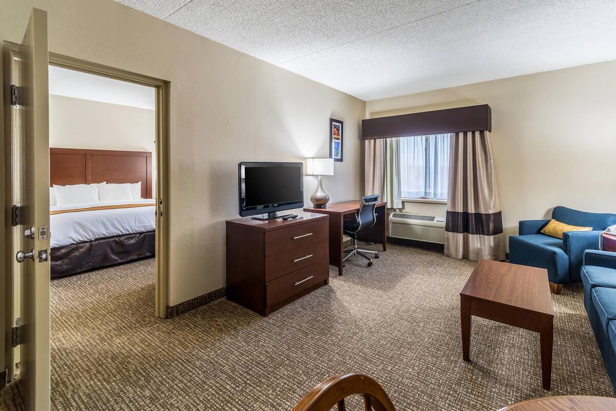 A hotel room with a bed, a desk with a TV, a chair, a sofa, and a window with curtains.