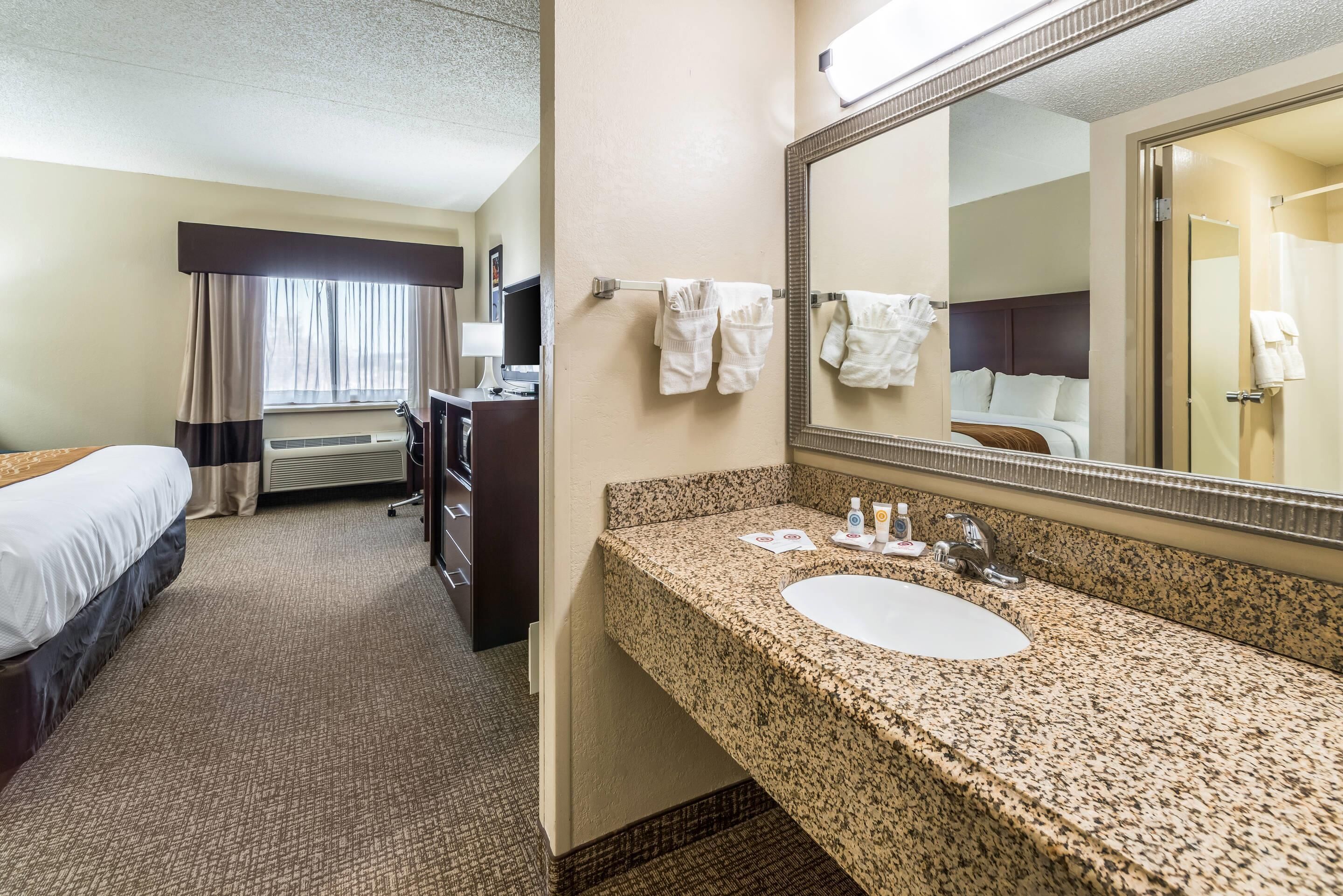 A hotel room with a bed, a desk, a bathroom with a sink and mirror, and a window with curtains.