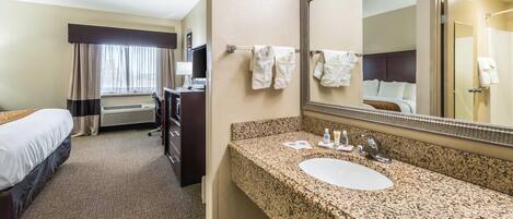 A hotel room with a bed, a desk, a bathroom with a sink and mirror, and a window with curtains.