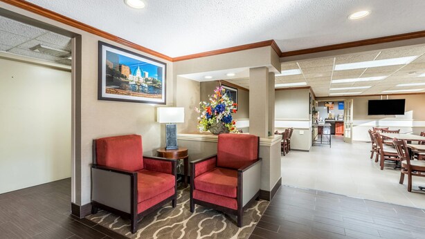 A hotel lobby with a reception desk, two red armchairs, a floral centerpiece, a framed cityscape picture, and a dining area in the background. - Comfort Inn Festus - St Louis South (Festus)