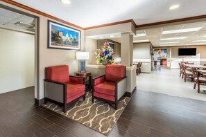 A hotel lobby with a reception desk, two red armchairs, a floral centerpiece, a framed cityscape picture, and a dining area in the background. - Comfort Inn Festus - St Louis South (Festus)