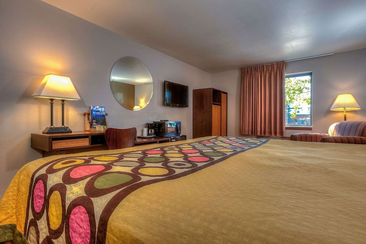 A hotel room with a large bed, a desk with a lamp, a mirror, a television, and a window with curtains.