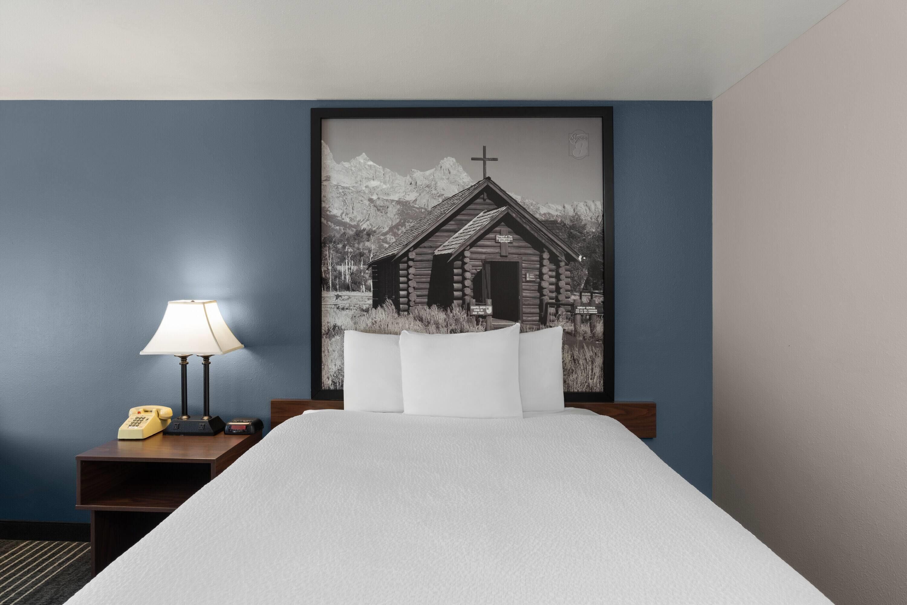 A hotel room with a bed, a nightstand with a lamp, and a framed picture of a mountain cabin.