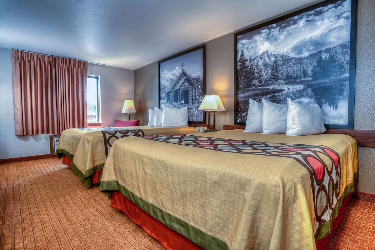 A hotel room with two beds, a large window with curtains, a lamp, and a painting of a mountain landscape.