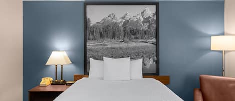 Una habitación de hotel con cama, una mesita de noche con lámpara, una silla y un cuadro grande con paisaje de montaña.
