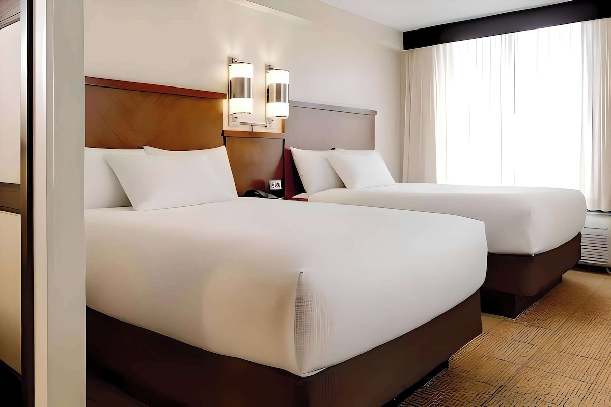 Ein Hotelzimmer mit zwei Betten, einem Holzkopfteil, Wandleuchten und einem großen Fenster mit Vorhängen.
