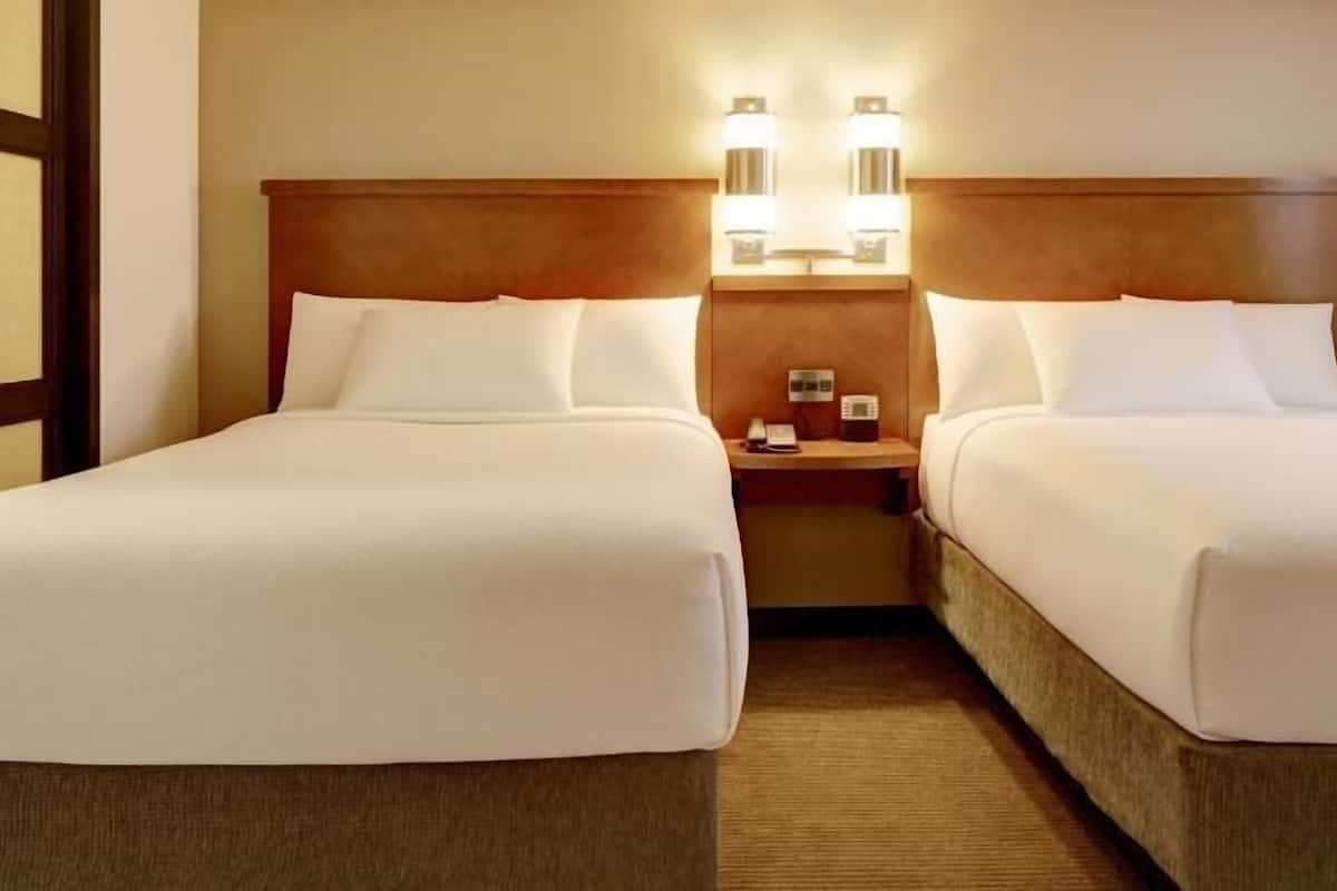 A hotel room with two beds, a nightstand with a phone and a lamp, and a wall-mounted light fixture.