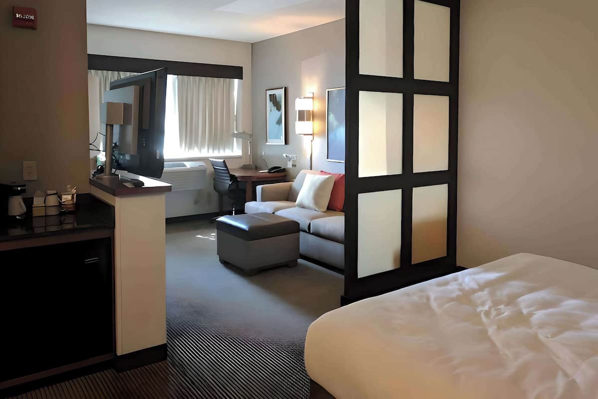 A hotel room with a bed, a desk with a computer, a sofa, and a large window with curtains.