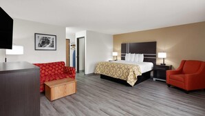 A hotel room with a bed, a red sofa, a wooden side table, a TV, and a nightstand with a lamp. - Baymont by Wyndham Green Bay (Green Bay)