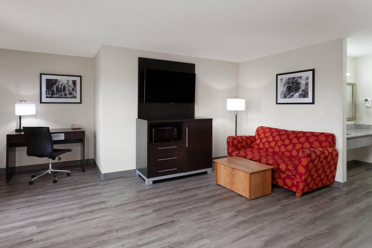 A modern hotel room with a wooden desk, a red patterned sofa, a flat-screen TV, and a small wooden side table.