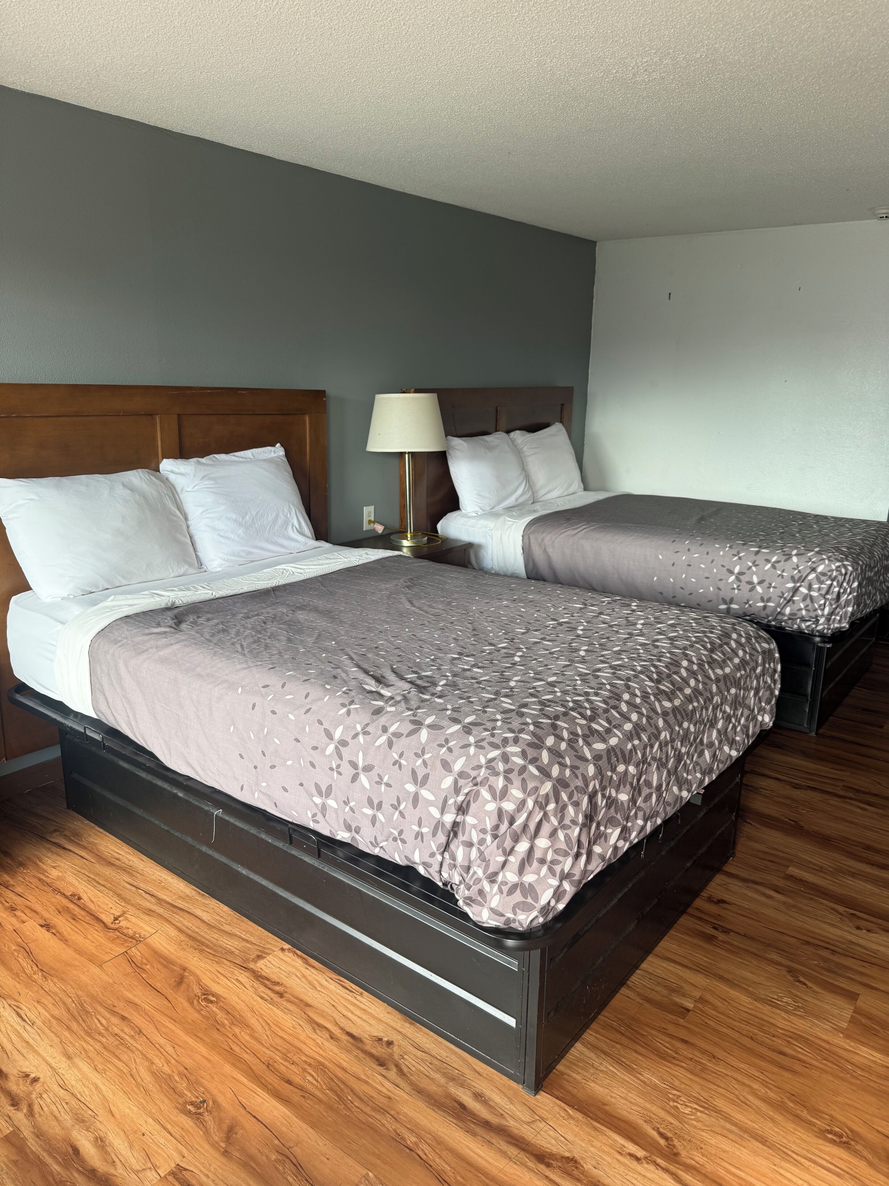 A hotel room with two beds, a nightstand with a lamp, and wooden flooring.