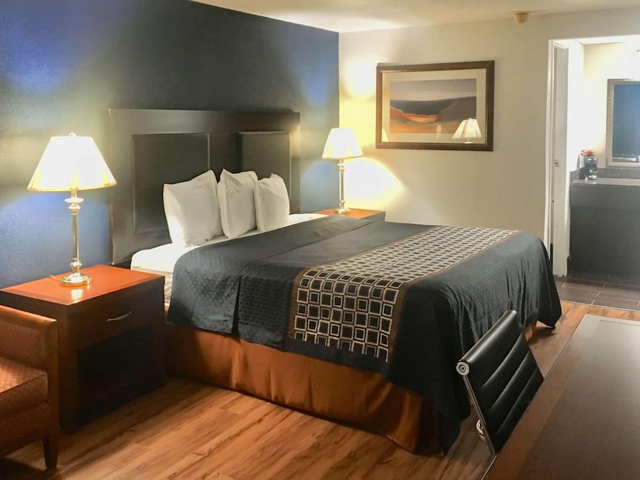 A hotel room with a bed, two bedside tables with lamps, a chair, a desk, a mirror, and a painting on the wall.