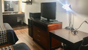 Et hotelværelse med seng, skrivebord med computer og skærm, skrivebordslampe, spejl og et lille tekøkken.