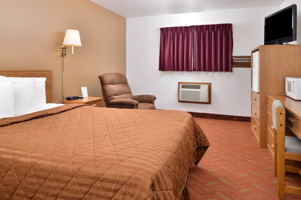 A hotel room with a large bed, a chair, a desk with a lamp, a television, and a microwave.