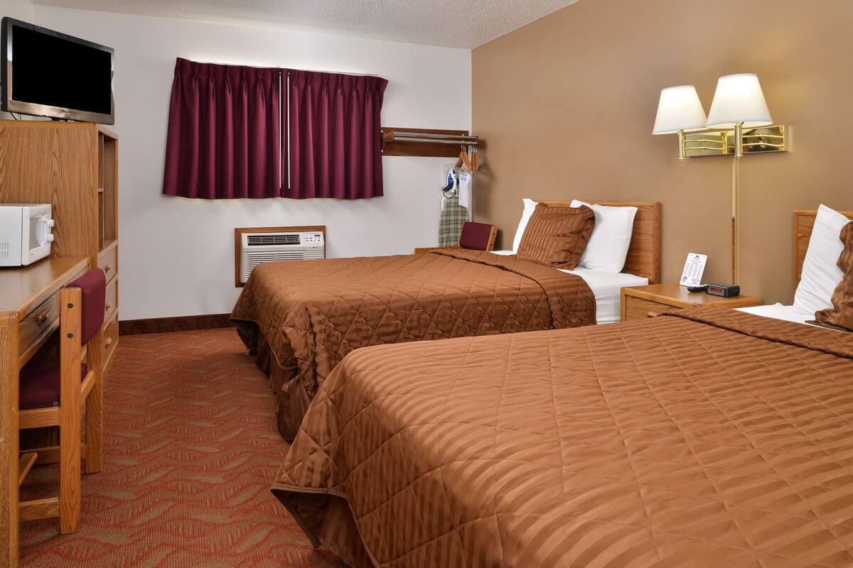 A hotel room with two beds, a TV, a desk with a chair, a wardrobe, a window with curtains, and a lamp.