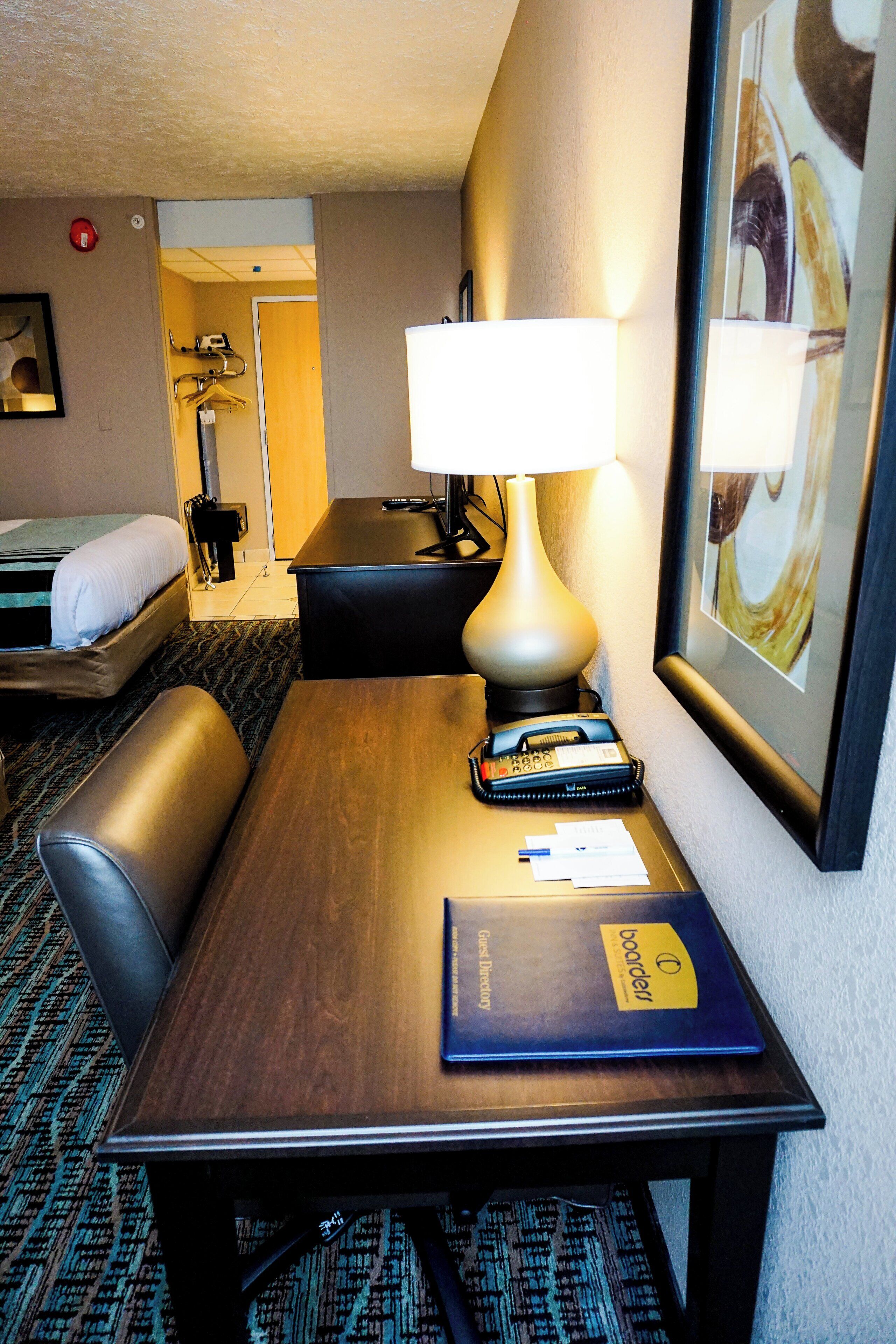 A hotel room with a wooden desk, a lamp, a bed, a chair, and a framed picture on the wall.