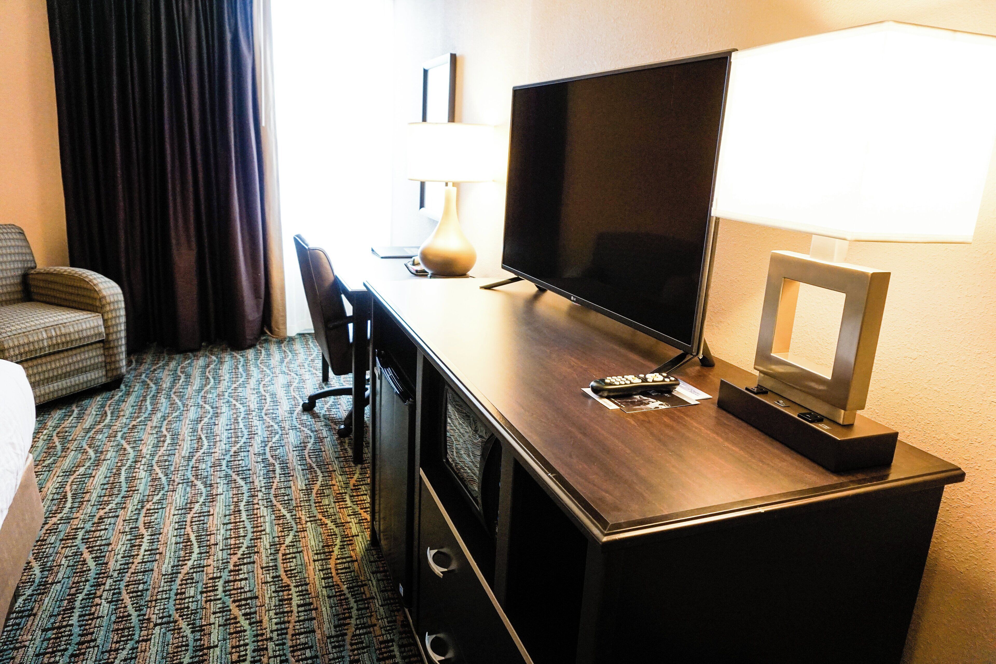 A hotel room with a large flat-screen TV, a desk with a lamp, a chair, and a bed.