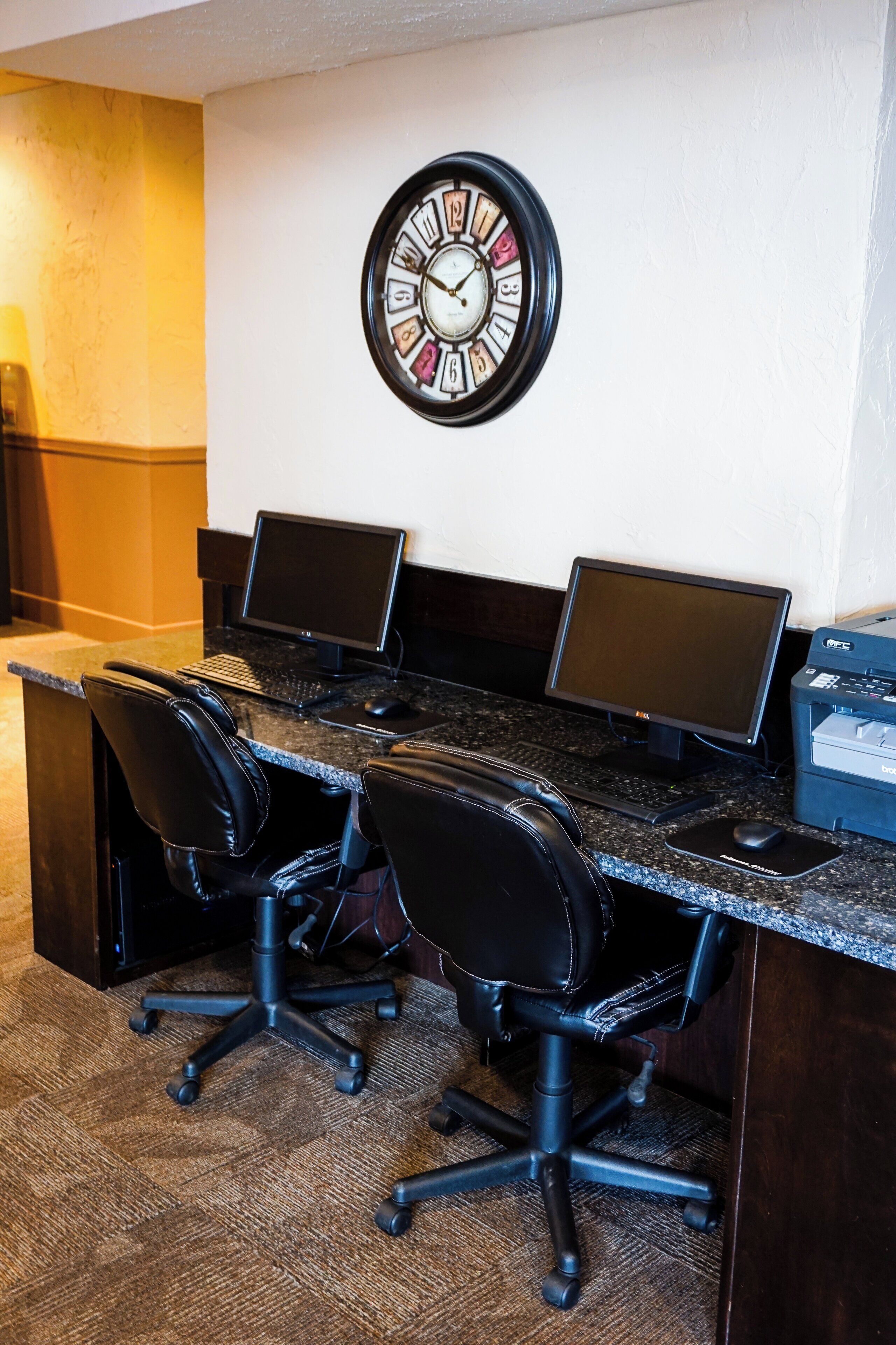 Een werkplek in een hotel met drie bureaus, twee stoelen, een klok en een printer.