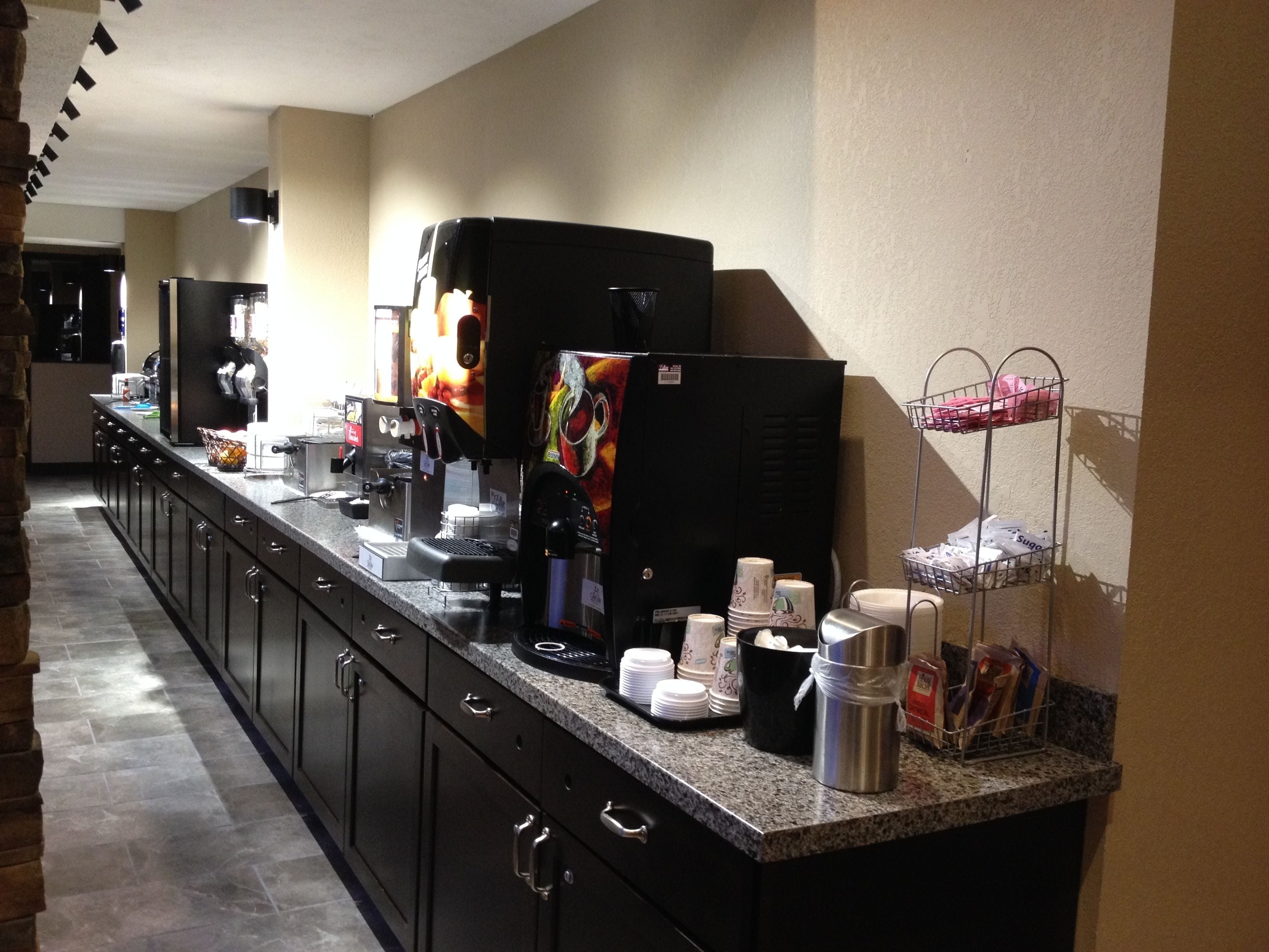 Un long comptoir abrite des machines à café, un réfrigérateur et un distributeur de boissons. On y trouve des piles de gobelets en papier et un présentoir métallique contenant des sachets.
