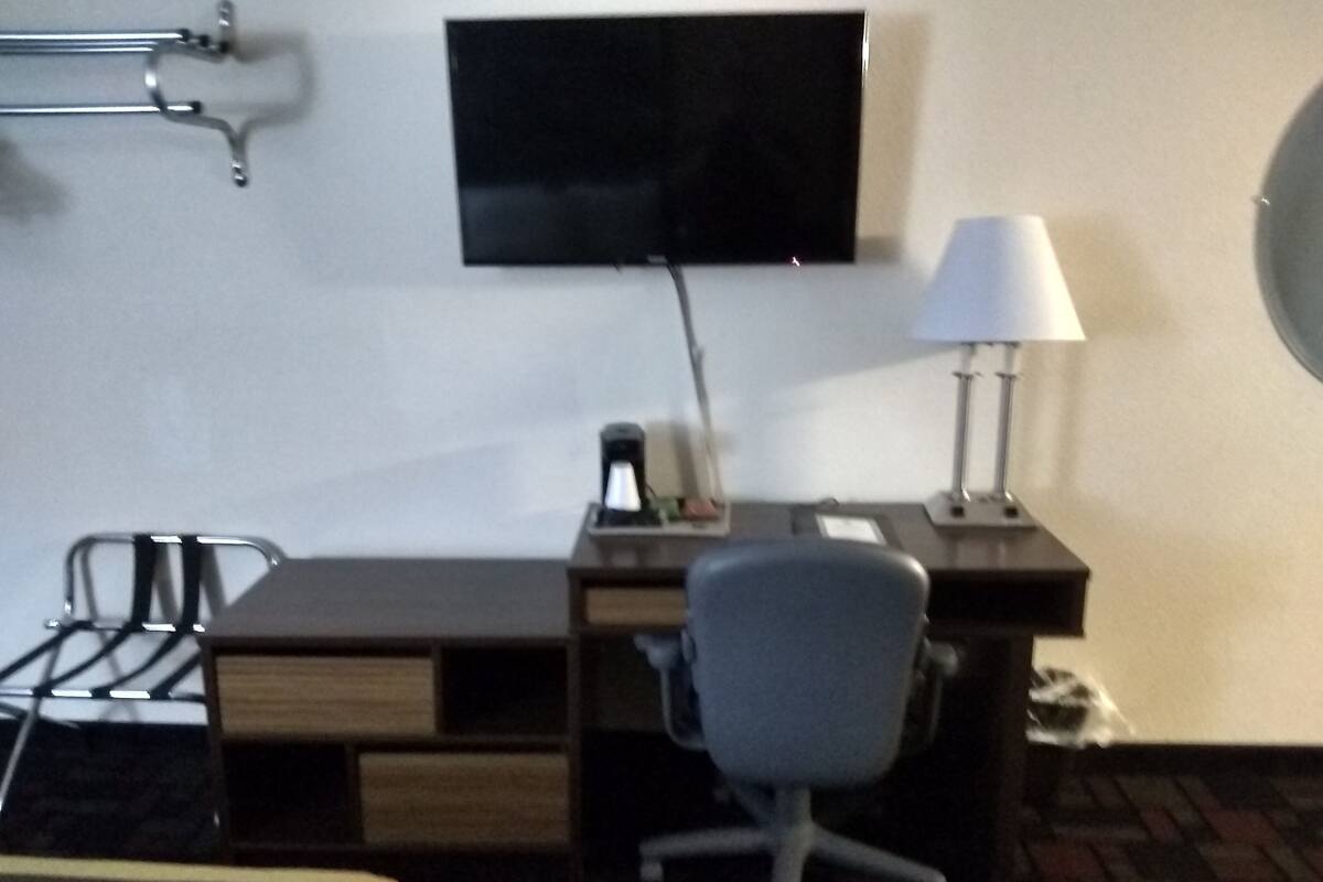 A hotel room with a bed, a desk, a chair, a TV mounted on the wall, and a lamp on the desk.