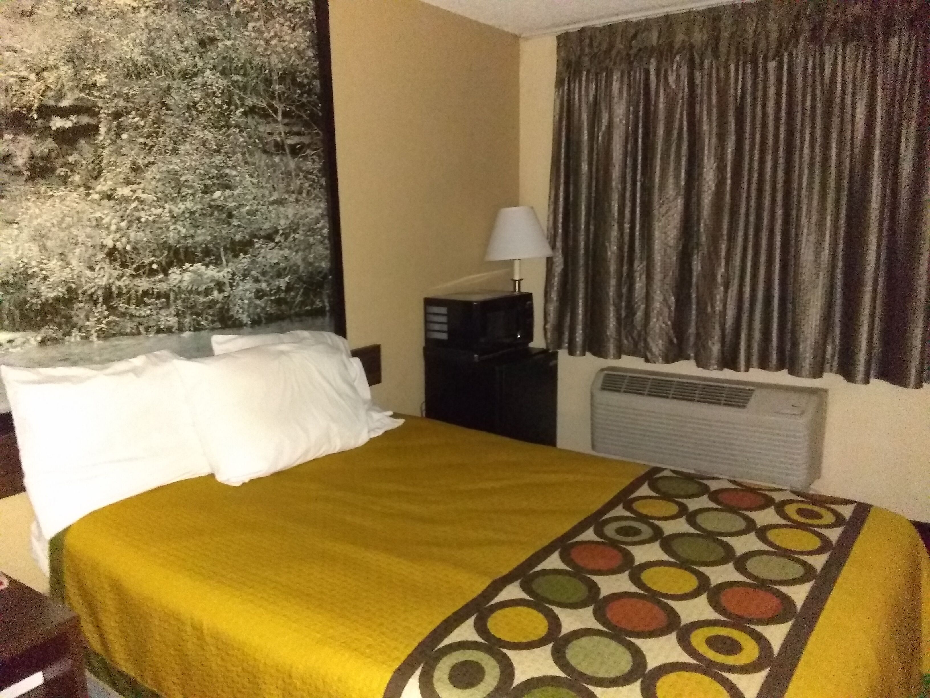 Habitación de hotel con una cama, una mesita de noche con lámpara, una ventana con cortinas y un aire acondicionado instalado en la pared.