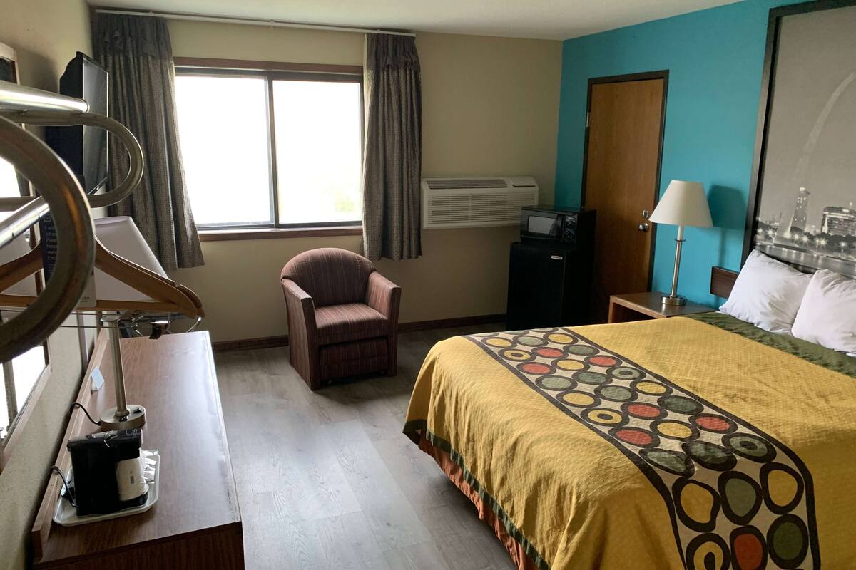 A hotel room with a bed, a chair, a desk with a computer, an air conditioner, and a window with curtains.