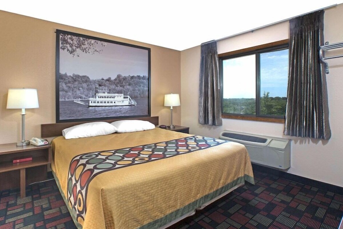 Une chambre d’hôtel avec un grand lit, deux lampes de chevet, une fenêtre avec des rideaux, un climatiseur fixé au mur et un grand tableau encadré représentant un bateau, accroché au mur.