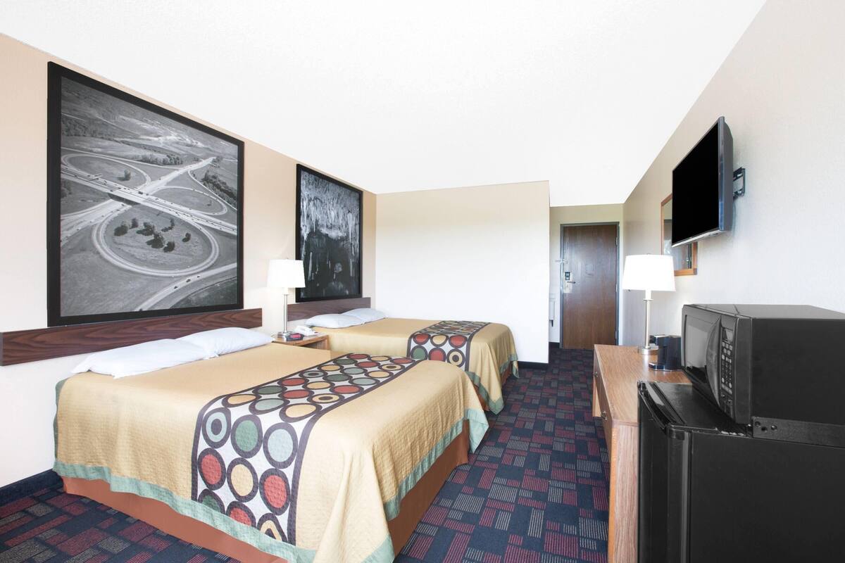 A hotel room with two beds, a television, a refrigerator, and two large paintings on the wall.