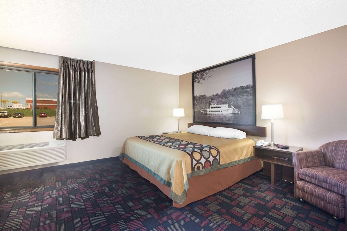 A hotel room with a bed, two bedside tables, a chair, a window with curtains, a wall-mounted air conditioner, and a painting of a boat on water.