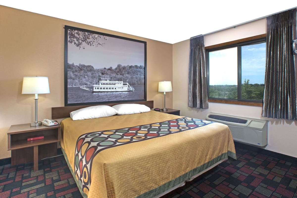 A hotel room with a bed, two bedside tables, a window with curtains, a wall-mounted air conditioner, and a large picture of a boat on the wall.