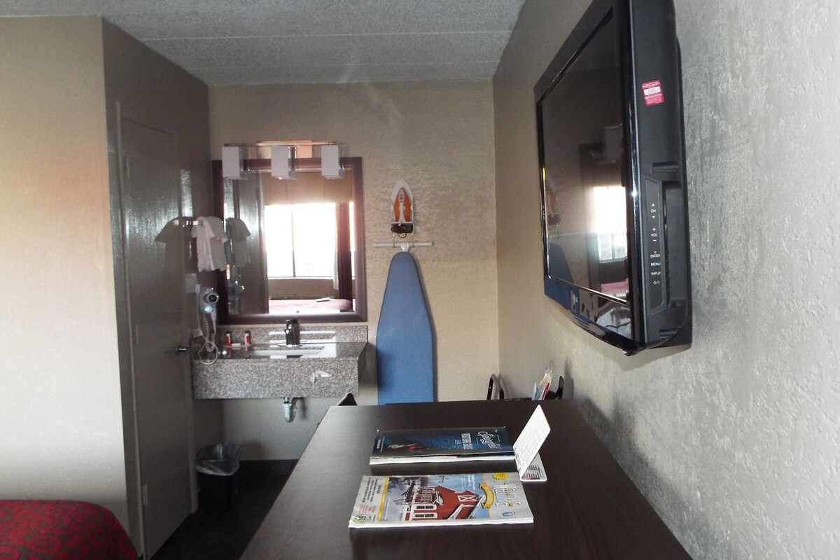 A hotel room with a TV, a small table with magazines, a blue chair, a sink, and a mirror.